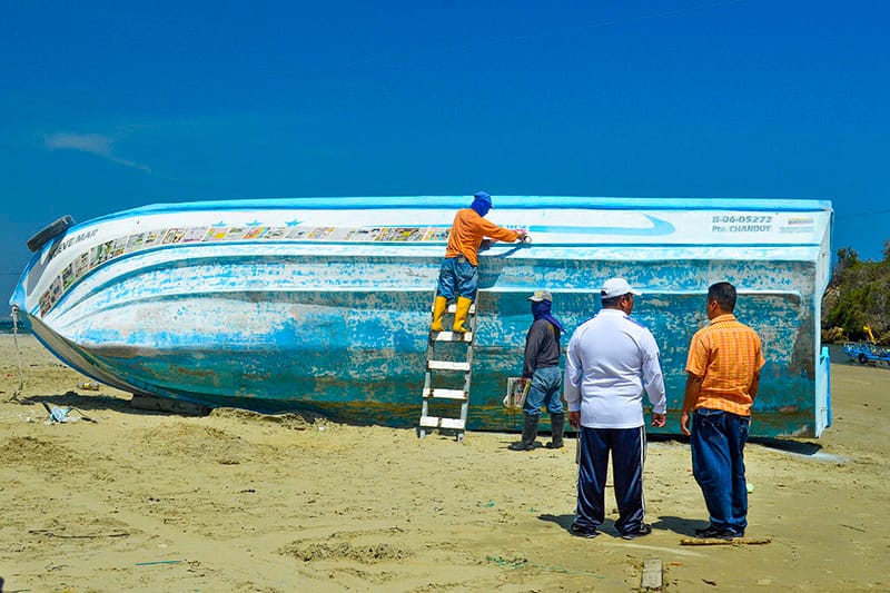 A man paints a blue and white fishing boat against a bright blue sky in Palmar, Ecuador