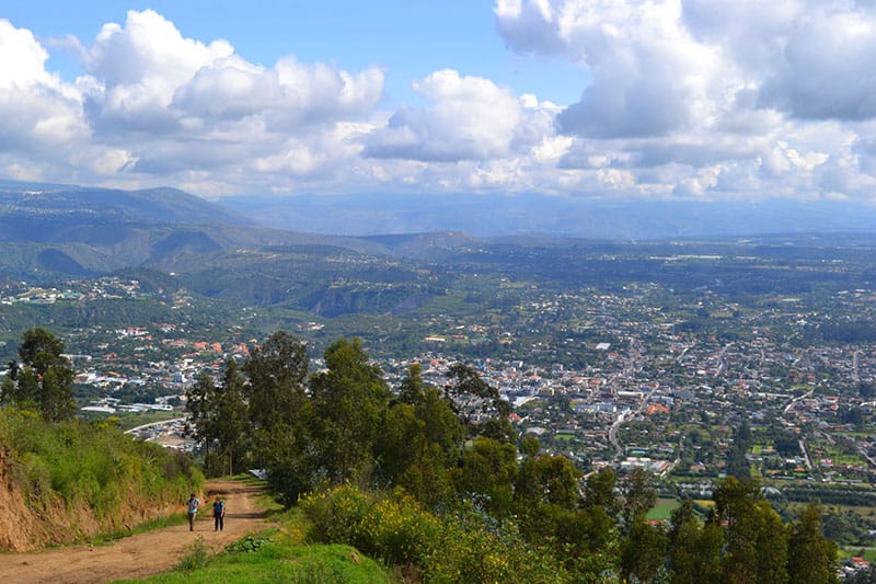 A wide shot showing Quito, Ecuador from the top of a hill with two people walking on a dirt path and puffy clouds in the distance