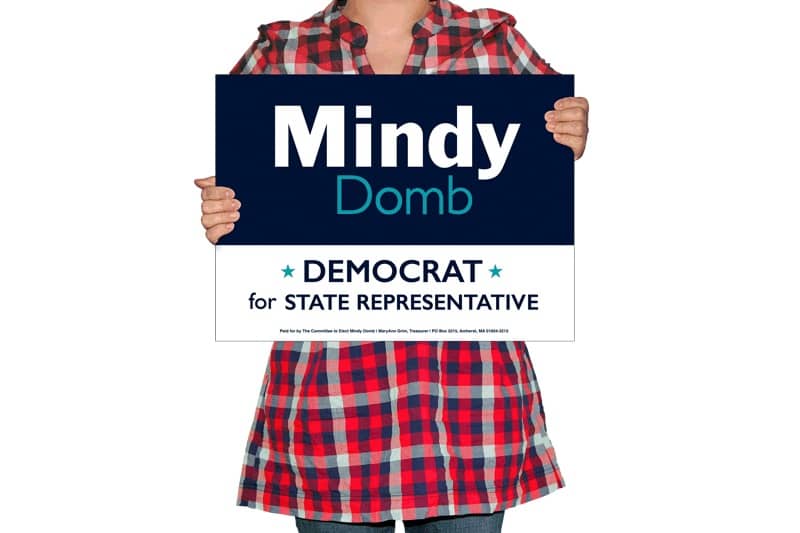 A woman wearing red plaid holds a lawn sign for Mindy Domb, state representative